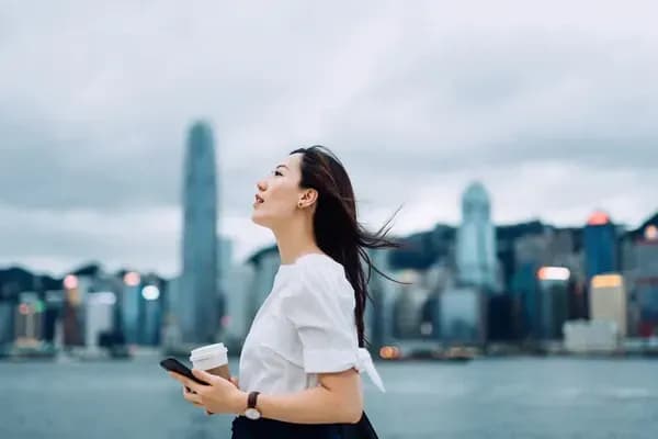 흰색 블라우스를 입은 여성이 커피와 스마트폰을 들고 도시의 해안가에서 하늘을 올려다보는 모습.